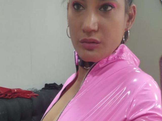 PaulaCamis with pink shiny latex jacket, zipped to chest, showing cleavage, hoop earrings, pink eyeshadow, and serious expression, standing in a room with a black couch.