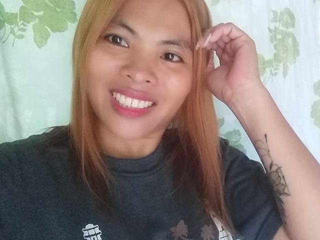 Chard69 has light brown skin, straight orange hair, and a tattoo on their right arm. They're smiling with pink lipstick, wearing a black shirt, and touching their hair.