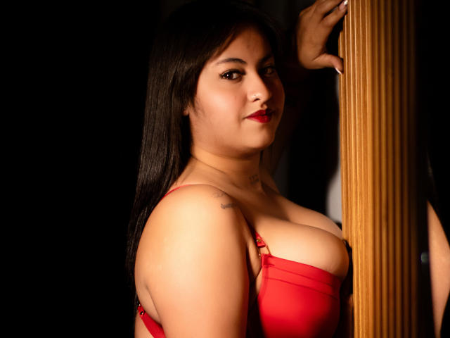 SharonParkerr with long black hair, red bra, and tattoo on shoulder, leaning against wooden pillar, looking at camera with red lipstick, showing her tits and slight smile.