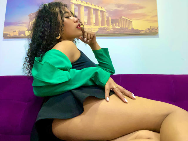 SophieSonyas with curly black hair, green off-shoulder top, black bottom, red lipstick, sitting on purple couch, hand on thigh, looking up, showing ass, white nail polish.