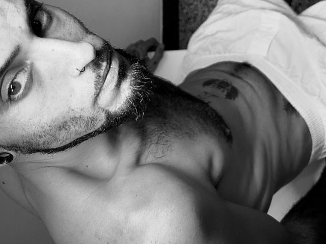 Black-and-white selfie of HardyGeorge with a nose ring, beard, and ear gauges, lying shirtless, exposing a tattoo on his chest, with white pants, showing a hairy chest.