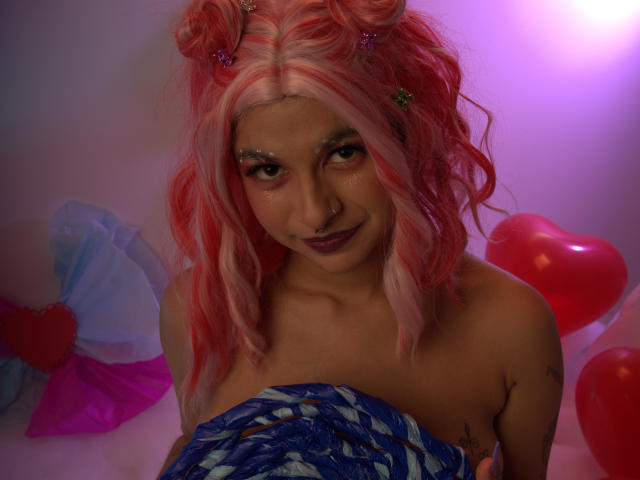 LaraAhB with pink and white wavy hair, glittery eyes, purple lipstick, topless, holding blue and white fabric, surrounded by red balloons and a pink heart.