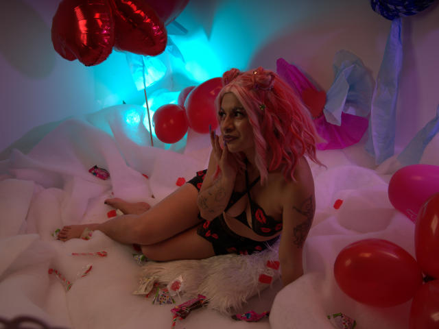 LaraAhB, avec ses cheveux roses, ses tatouages, en lingerie noire à bretelles, assise sur un lit blanc moelleux avec des ballons rouges, des guirlandes de bonbons et un coussin en fausse fourrure blanche, regardant l'appareil photo..