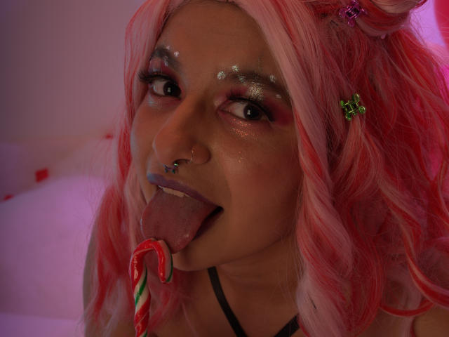 LaraAhB, avec ses cheveux roses, son anneau de nez et sa pince à cheveux verte en forme d'étoile, tenant une sucette dans sa bouche, la langue dehors, les yeux scintillants et le rouge à lèvres violet, dans une pose sexy..