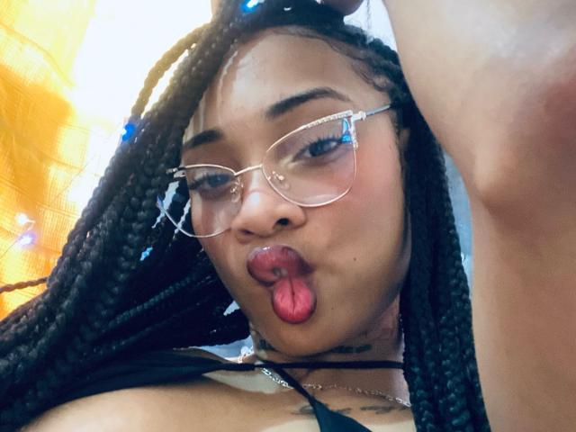 ElaHott with braided black hair, gold-rimmed glasses, and her tongue out, showing red lips. She's wearing a black top, and her small tits are partially visible.
