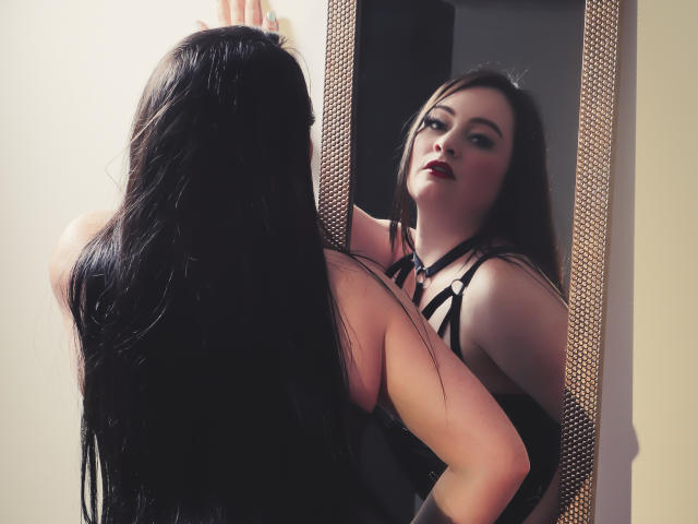 KristelRouse with long black hair, staring at herself in a mirror, wearing a black strappy top, her reflection shows red lipstick, her hand on the mirror.