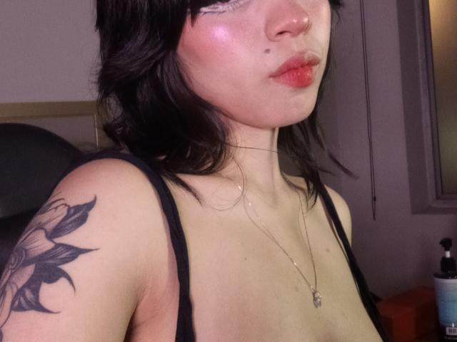 Natasha Hottson mit schwarzen Haaren, glänzenden rosafarbenen Lippen, einem Nasenring und einem Tattoo an der rechten Schulter, trägt ein schwarzes Oberteil, das ihre Brüste zeigt, und eine Halskette in einem schlecht beleuchteten Raum..