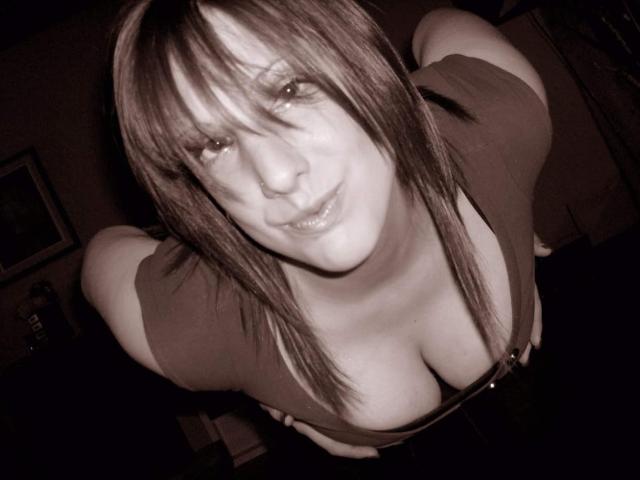 ValentinaNataliia69Hot with medium-length brown hair, looking down, smiling, wearing a low-cut top showing large, visible breasts, in a dimly lit, sepia-toned photograph.