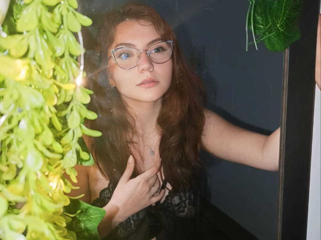 ArabellaShine with long red hair, glasses, and black lace dress, hand on chest, green leaves to the left, looking slightly bored. Boobs visible through dress.