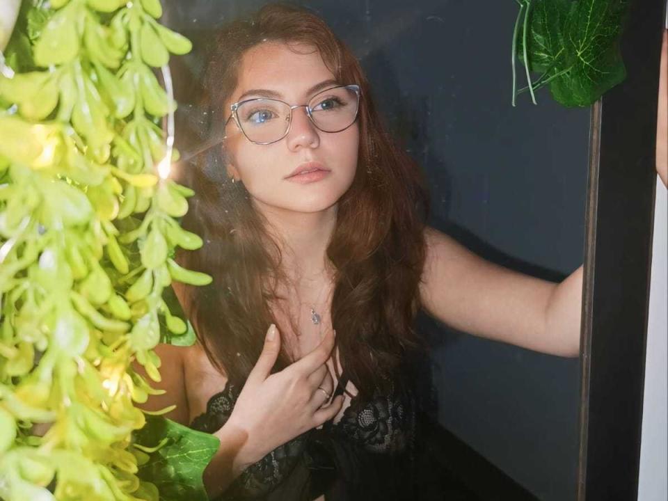 ArabellaShine with long red hair, glasses, and black lace dress, hand on chest, green leaves to the left, looking slightly bored. Boobs visible through dress.