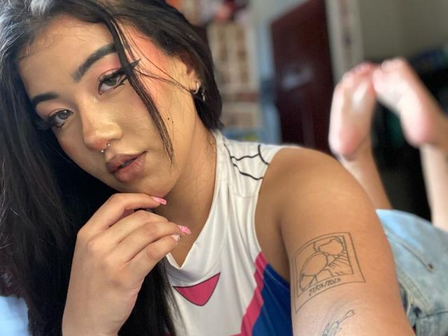 KelseyCoob has dark hair, a nose ring, pink eyeliner, and a tattoo on her arm. She's wearing a white sleeveless top, resting her chin on her hand.
