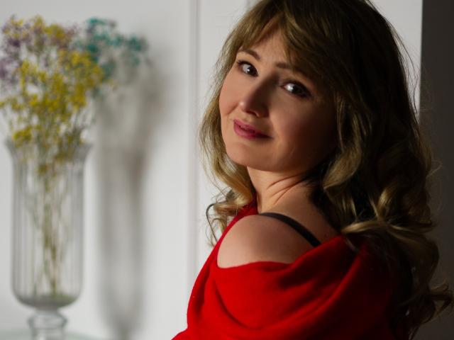 JustineFlirt with wavy brown hair, red off-shoulder top, black strap visible, smiling at camera, soft light, subtle makeup, gentle expression, blurred flowers in background, relaxed, sexy.