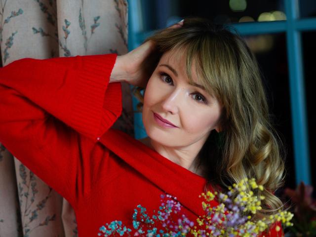 Justine, com cabelo loiro, suéter vermelho e pequenas flores em primeiro plano, sorrindo gentilmente com uma mão no cabelo, mostrando seu rosto adorável e atraente e seus lábios rosados delicados..