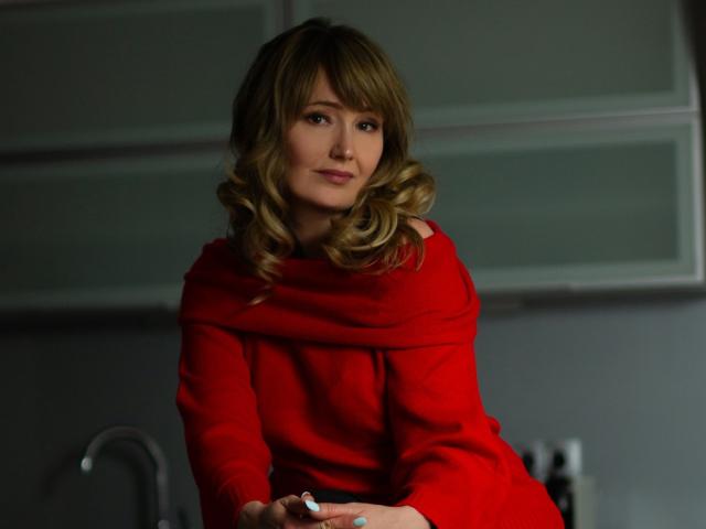 JustineFlirt with wavy blonde hair, wearing a red off-shoulder sweater, sitting in a kitchen, looking slightly to the side with a subtle smile, holding a white object.
