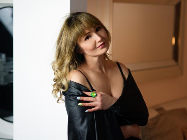 JustineFlirt with blonde curls, black off-shoulder top, green ring, and blue nails, showing her tits, leaning against a white wall, looking sexy, in a soft light.