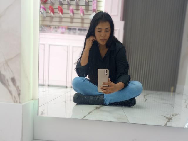 ArianithaXox sitzt mit angelegten Beinen auf einem glänzenden Boden, trägt ein schwarzes Hemd und blaue Jeans und hält ein goldenes Telefon, auf das sie mit einem leicht schmollenden Gesicht schaut..
