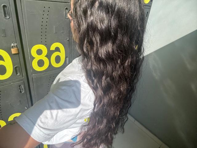 AlisonTatcher's long, wavy dark brown hair, back to the camera, sunlight highlighting her white t-shirt, grey lockers with yellow numbers in the background.