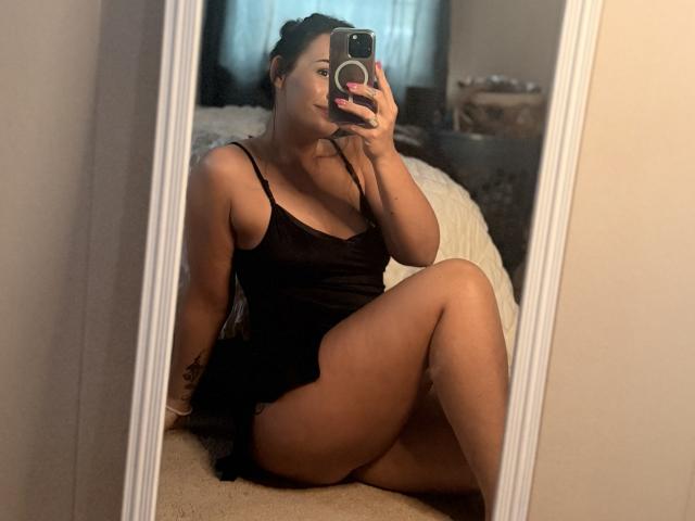 MaggieJane is sitting in front of a mirror, wearing a black tank top and shorts, showing her tits and ass, with a phone in her hand, taking a selfie.