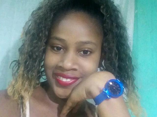 OliviaHot69 with curly brown hair, medium brown skin, red lipstick, smiling, wearing a blue wristwatch, pointing at camera. Boobs not visible. Ass not visible. Pussy not visible.