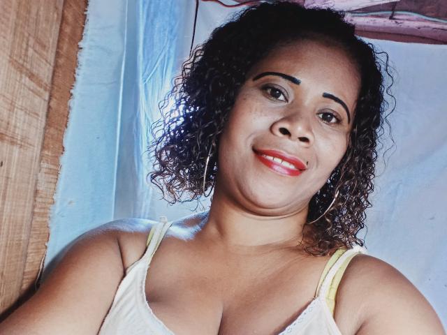 Emie69 has curly brown hair, brown eyes, red lipstick, and is smiling with her tits slightly visible in a white tank top, wearing large hoop earrings.