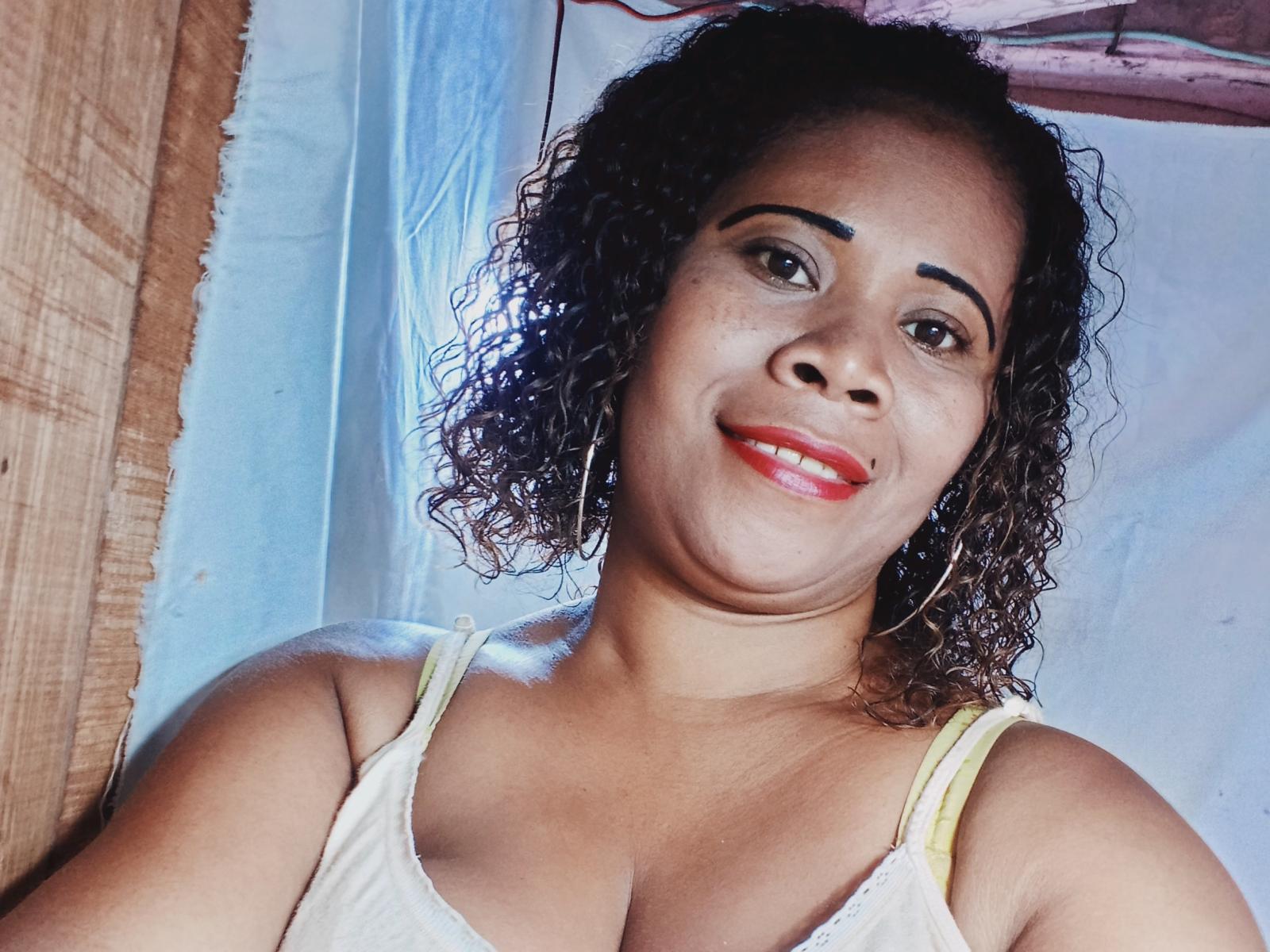 Emie69 has curly brown hair, brown eyes, red lipstick, and is smiling with her tits slightly visible in a white tank top, wearing large hoop earrings.