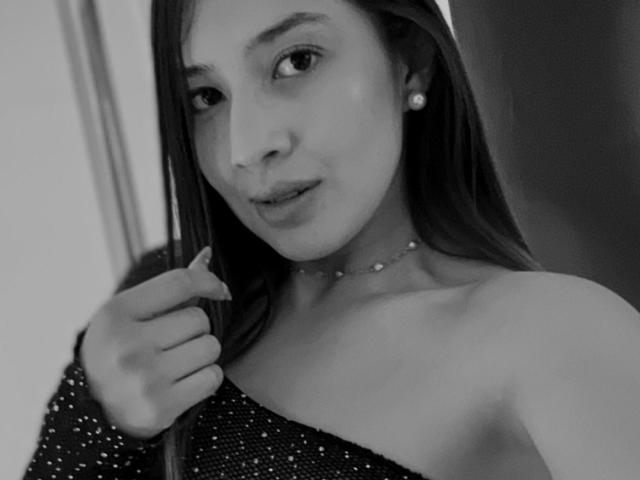 Emma Lombardi, met lang donker haar, kleine parel oorbellen, een glinsterende jurk met een open schouder, die de jurk vasthoudt met haar linkerhand, een zwart-wit selfie, met een lichte glimlach, kijkend naar de camera..