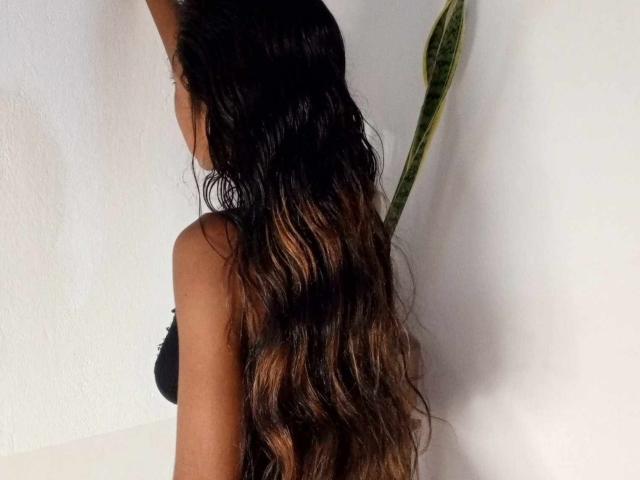 AshaSadha's long, wavy brown hair with blonde highlights touches a white wall, her black bra-clad back visible, a green plant partially in frame on the right.