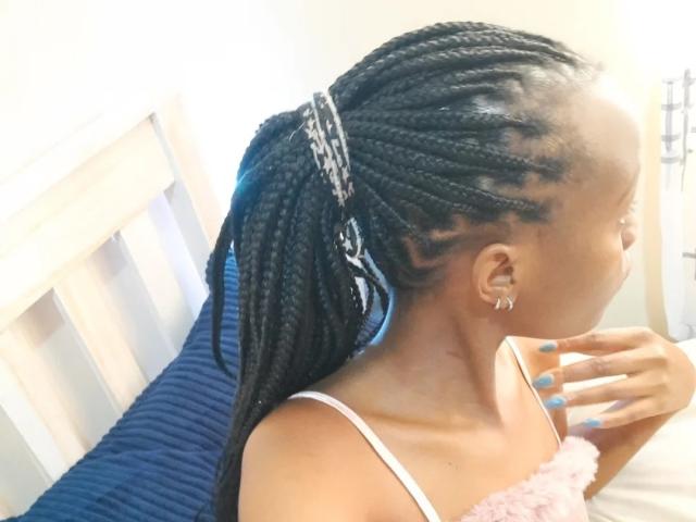 EbonySwt met lange zwarte vlechten, witte beha, roze pluizige top, blauwe nagels, kleine oorringen, profiel van de zijkant, wegkijkend, hand in de buurt van de mond, wit bedframe, blauw kussen..
