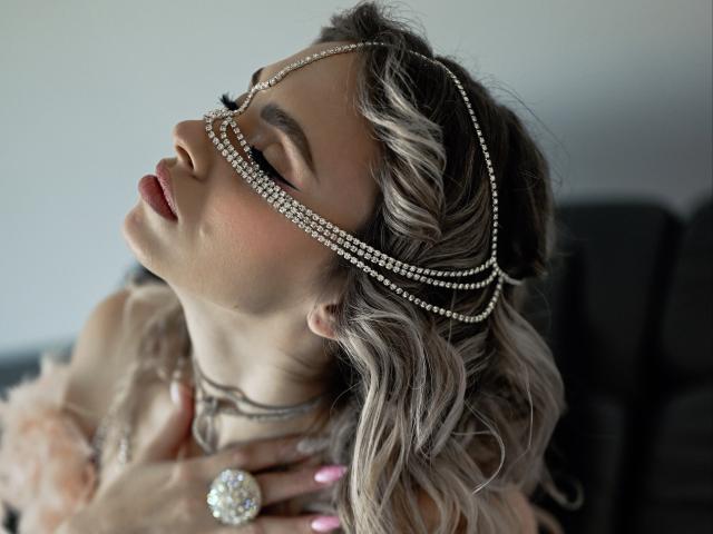 ChloeBright with wavy blonde hair, eyes closed, wearing a diamond chain headpiece, pink nails, and a large ring, head tilted back, hand on neck, soft makeup.