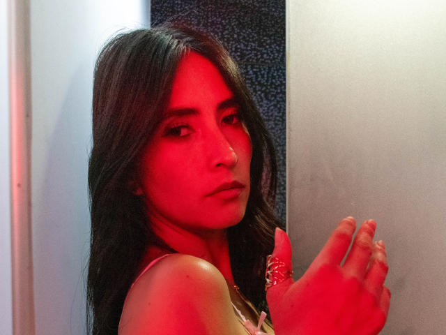JainaRogers with red-tinged skin, long dark hair, and a nose ring, hands up, wearing a pink top, looking at the camera with a serious expression.