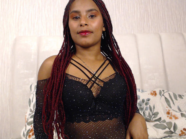 Samantha Roussee, con lunghe trecce rosse, indossa una top nera, trasparente e con spalline sottili, ricoperta di paillettes, che mette in mostra il suo seno piccolo e formoso e il rossetto rosso, ed è in piedi di fronte a un letto..
