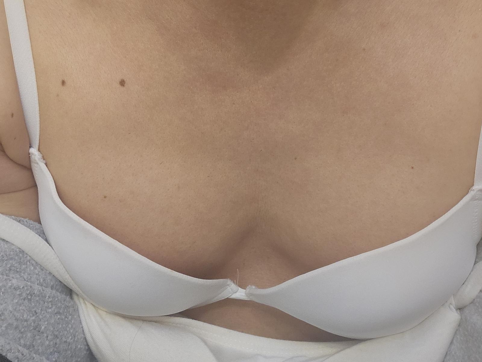 Andykiss's tits in a white bra, showing cleavage with a small mole on the right side. Andykiss's light skin is slightly textured. Grey garment partially visible.