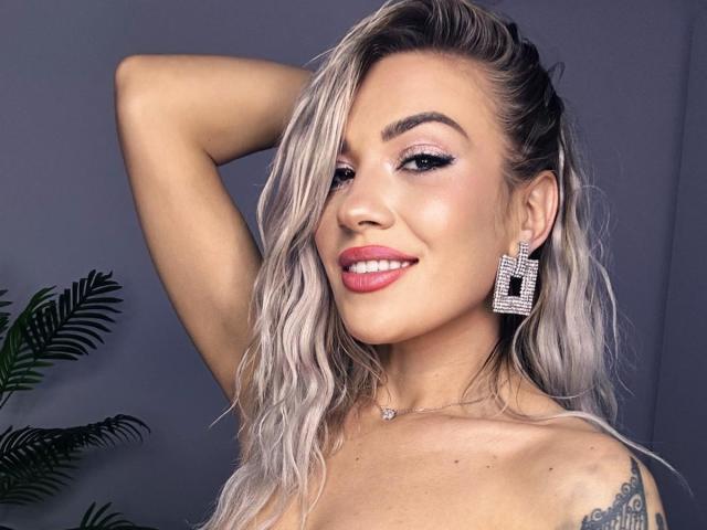 ZoeyMarie with blonde wavy hair, pink lipstick, and diamond earrings, smiling with one arm behind her head, showing a tattoo on her right shoulder. Boobs visible.