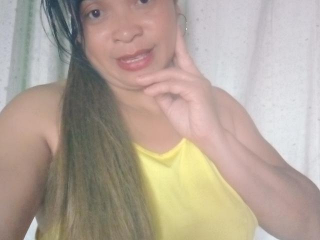 Rahel69 with long brown hair, yellow tank top, and hoop earrings, smiling, touching her cheek, showing small tits, and wearing subtle makeup in a casual selfie.
