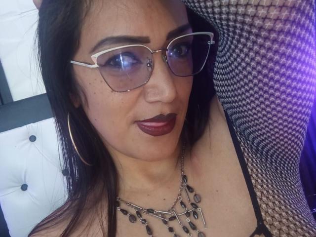 DayanaMarians69, con i capelli scuri, gli occhiali e il rossetto viola, indossa una maglietta nera, una collana a catena e orecchini a cerchio, guarda verso la fotocamera, con una luce viola sulla destra..