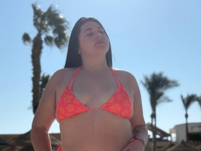 ArianaDarko with eyes closed, wearing a pink-orange floral bikini top, showing her tits, standing outdoors, palm trees in the background, sunlight on her dark hair.