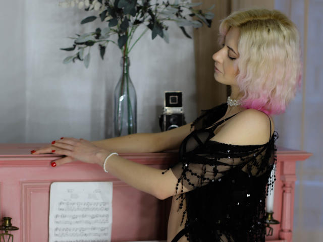 MiaBura with pink-to-blond hair, wearing black sequined top, hands on pink fireplace, red nails, white bracelet, camera on fireplace, vase with white flowers, side profile, sexy.