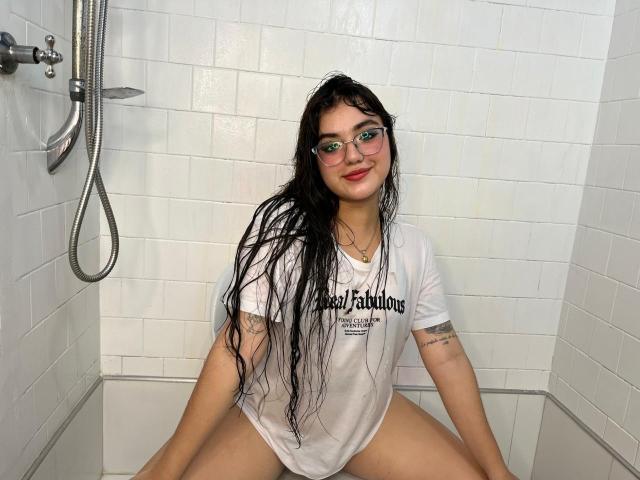 VictoriaSaens, con i capelli neri bagnati, occhiali verdi, camicia bianca e tatuaggi visibili, seduta in una doccia con piastrelle bianche, gambe divaricate, sorridente, senza pantaloni, che mostra la sua vulva..