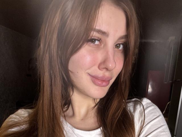 SensualMila has light brown hair, a cute nose, and full pink lips. She's smiling slightly with a flushed face, wearing a white shirt, in a dimly lit room.