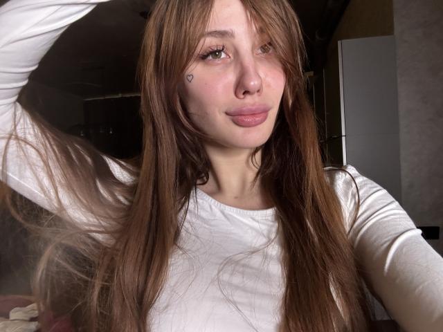 SensualMila mit langen, braunen Haaren, einem weißen, langärmeligen Hemd und einem herzförmigen Nasen-Tattoo, leicht geröteten Lippen, grünen Augen und einer Hand im Haar, sieht sexy und süß aus..