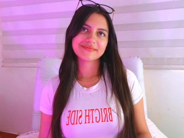 MiaLeiya with long brown hair, glasses on head, white shirt, red text, smiling, pinkish-purple lighting, white chair, sitting, medium breasts, cute, light skin, looking to the side.
