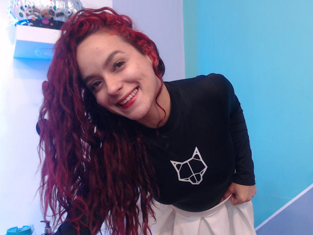 MaiaPark with red curly hair, smiling, wearing a black long-sleeve shirt with a white cat graphic and a white skirt, bending forward slightly. Her red lips match her hair.