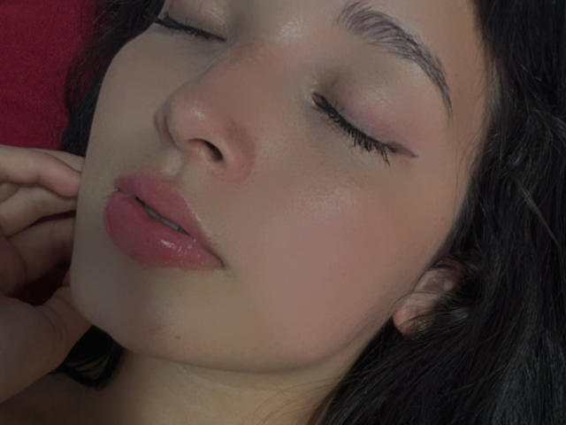 O rosto de Sarah Good, olhos fechados, batom rosa, pele brilhante, sobrancelhas pretas, cabelo preto, lábios rosados ligeiramente entreabertos, mão perto da bochecha, luz suave, fundo vermelho, expressão relaxada..