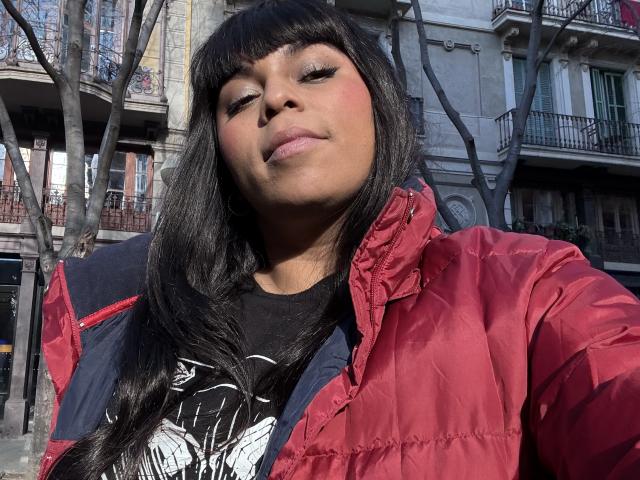 Chile con piel oscura y cabello negro, vistiendo una chaqueta roja hinchada y una camiseta negra, mirando orgulloso en un entorno urbano.