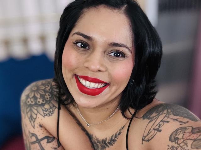 Chili mit schwarzen, schulterlangen Haaren, rotem Lippenstift und Tattoos lächelt in die Kamera und zeigt strahlend weiße Zähne brust und Dekolleté.