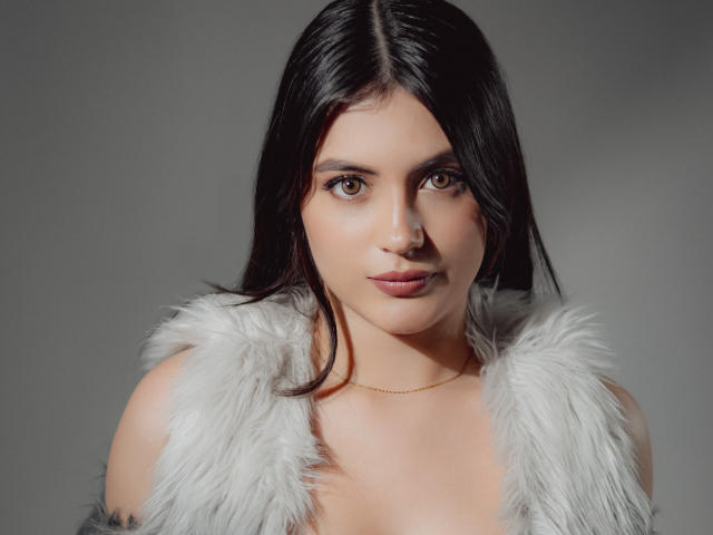 MiaLeiya with dark, straight hair, brown eyes, and a subtle smile. She's wearing a white, furry vest and a thin gold necklace. Boobs partially visible. Gray background.