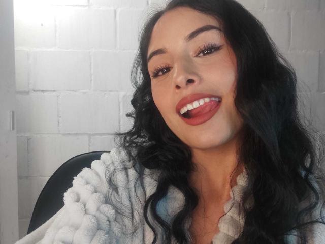 EmilyVv with long black wavy hair, brown eyes, and red lipstick, tongue out, smiling, wearing a white fluffy robe, sitting on a black chair.