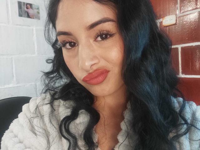 EmilyVv heeft zwart, golvend haar, bruine ogen en rode lippenstift, en draagt een witte, pluizige jas, met een subtiele glimlach. Haar borsten zijn niet zichtbaar, alleen haar gezicht..