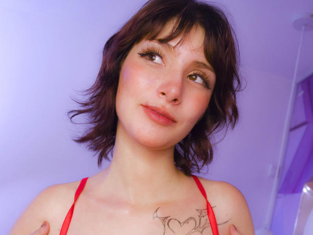 IsabellaHallad with short brown hair, eyeliner, and red top, looking sideways with a slight smile, showing tattoos on her chest and arms, in a soft purple-lit room.