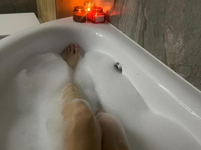 TianaStar's legs in a white bathtub with foamy water, her feet with pink toenails, and two red-lit candles on the edge. Her pussy is partially submerged.